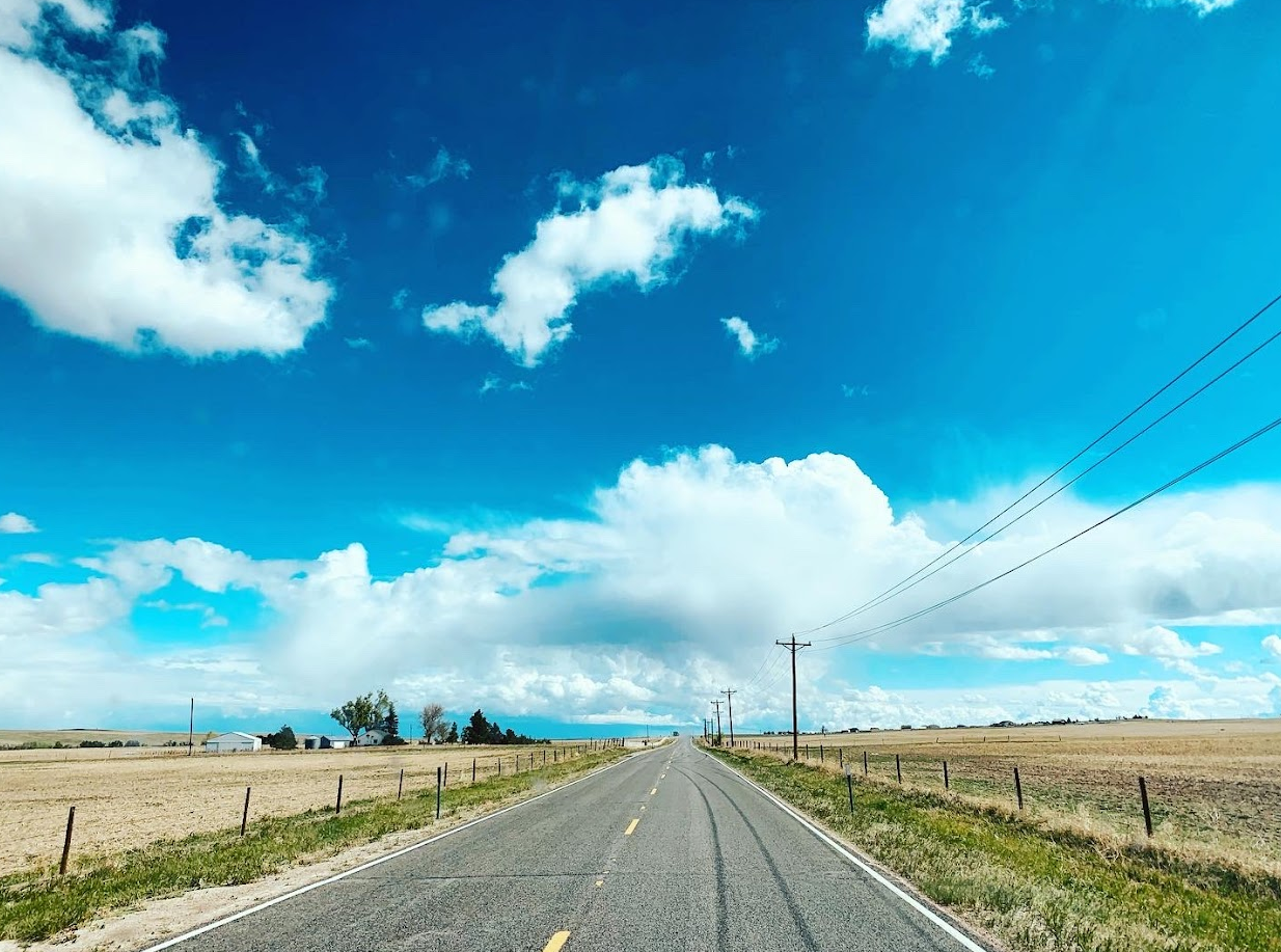 2 lane highway flanked by farms and covered with clouds against a blue sky